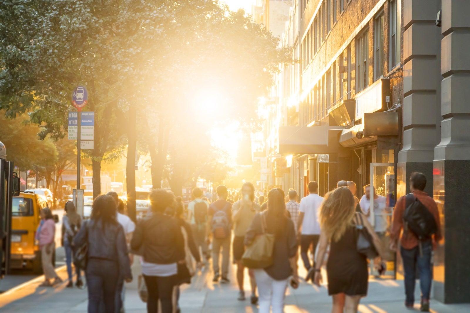 Man stands in the middle of a busy sidewalk looking at his cell phone while crowds of people walk around on 14th Street in Manhattan, New York City with the glow of sunlight in the background.