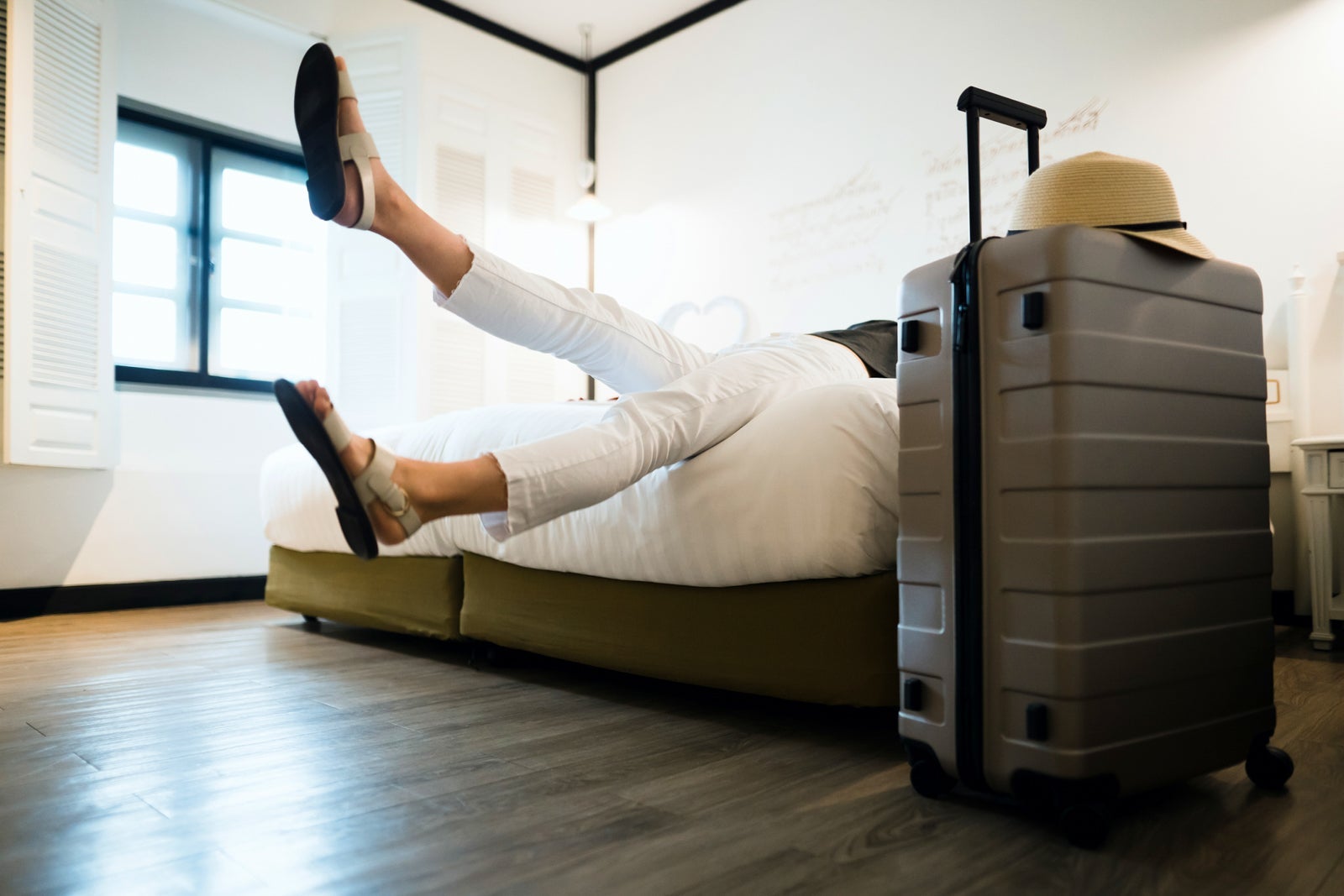 Young female traveller relaxing on bed in a hotel room, with a suitcase and straw hat by the bed