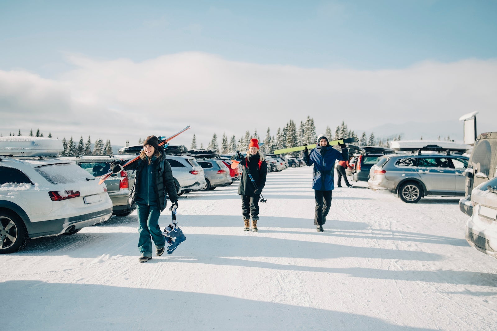 Female and male friends holding skis while walking on snow at parking lot against sky