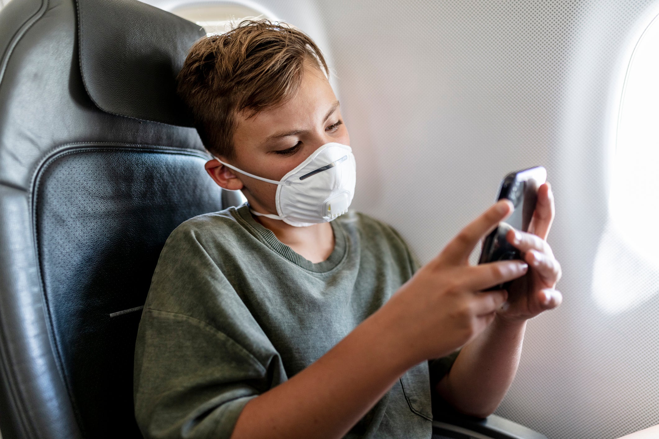 teenager boy is wearing his FFP3 protective mask while he is playing with his mobile phone in airplane