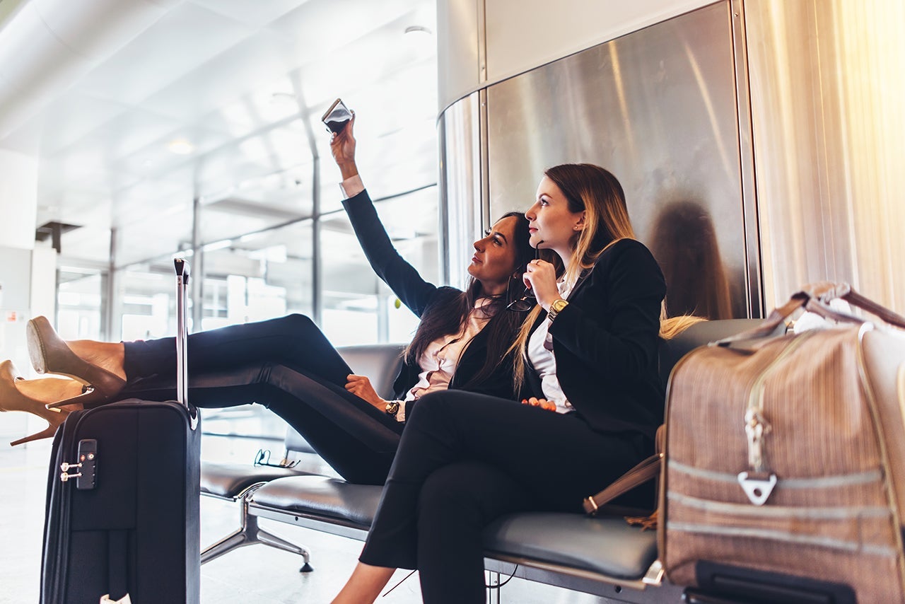 Two women talking selfie while waiting for a delay flight sitting with suitcases in airport terminal