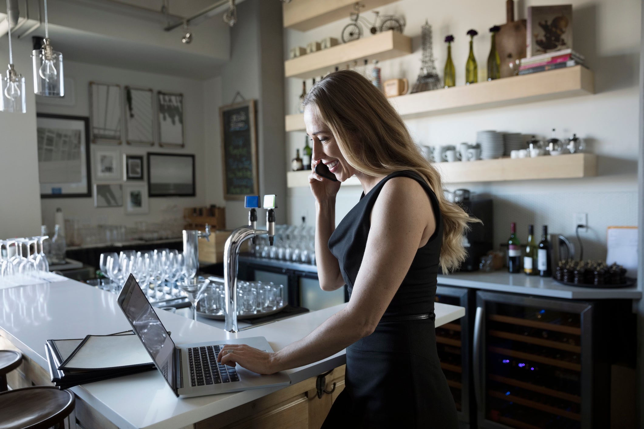 Female small business owner talking on smart phone and working at laptop in cafe