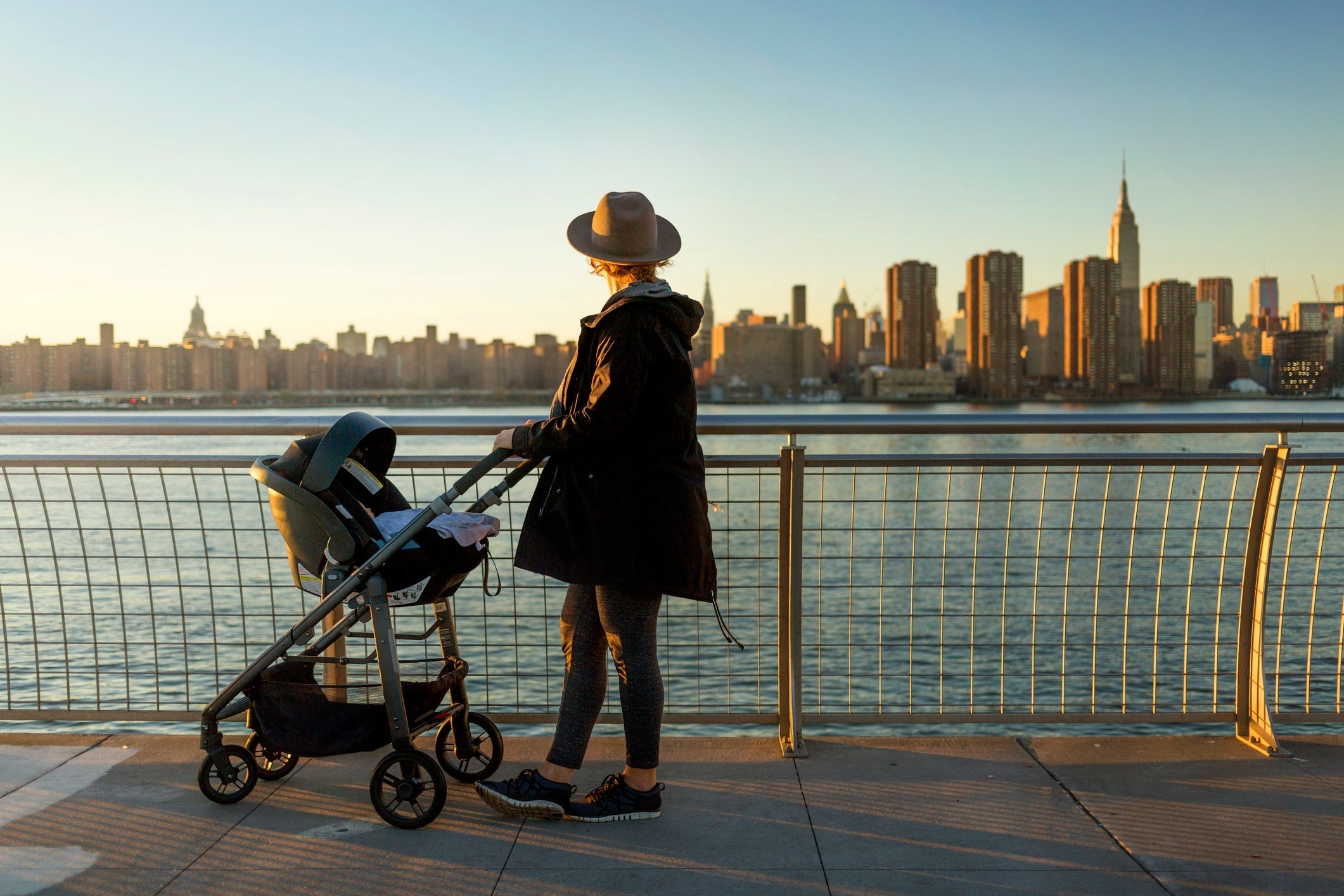 Side view of woman looking at city skyline while standing on promenade with baby carriage during sunset