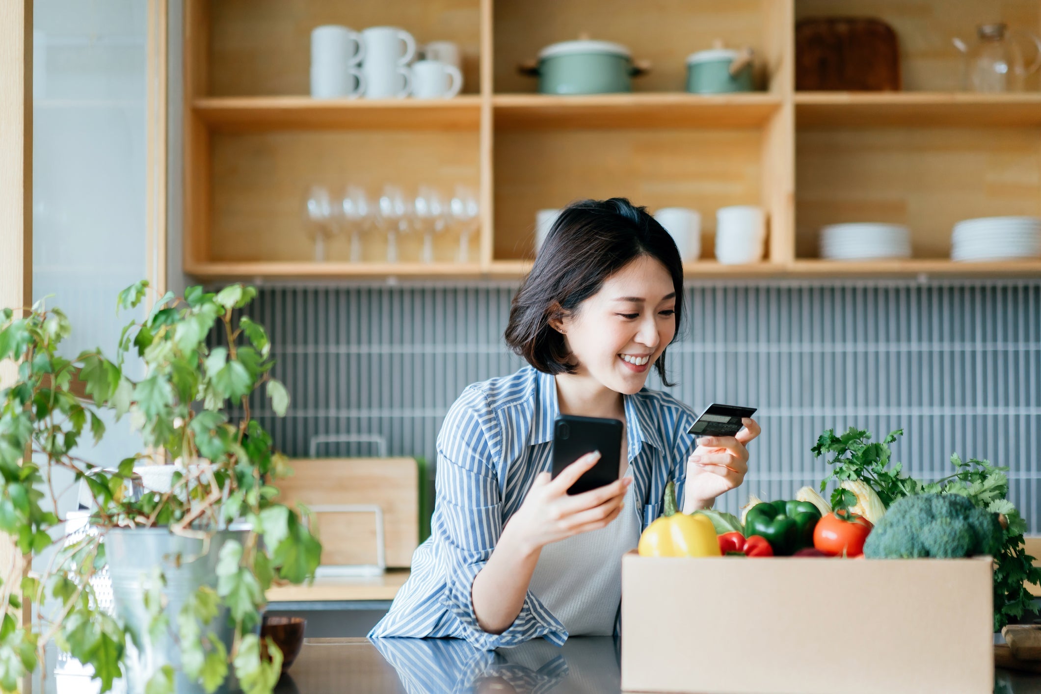 Beautiful smiling young Asian woman grocery shopping online with mobile app device on smartphone and making online payment with her credit card, with a box of colourful and fresh organic groceries on the kitchen counter at home