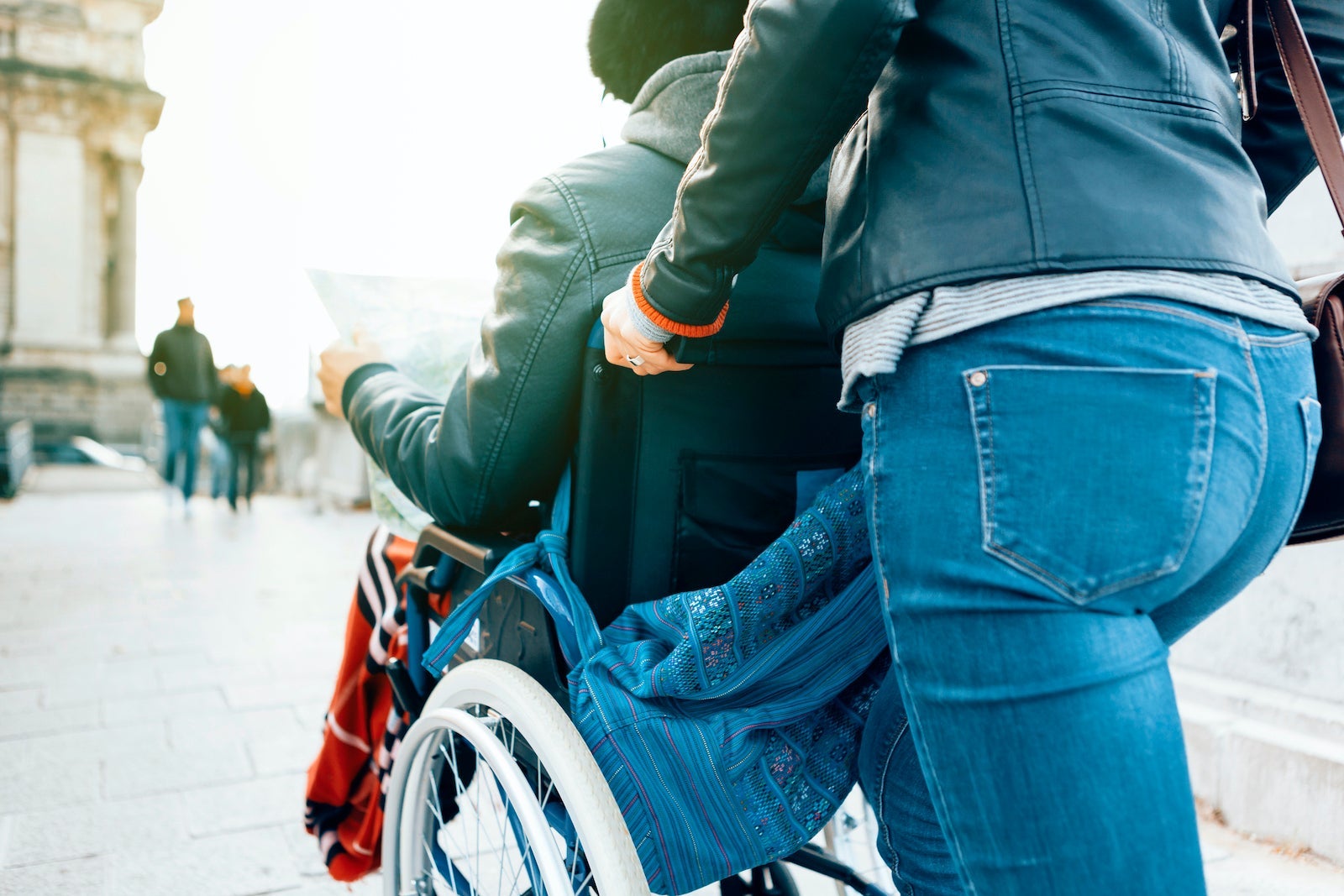Side View Of A Man Sitting In A Wheelchair While A Female Person Is Pushing Him Around The City