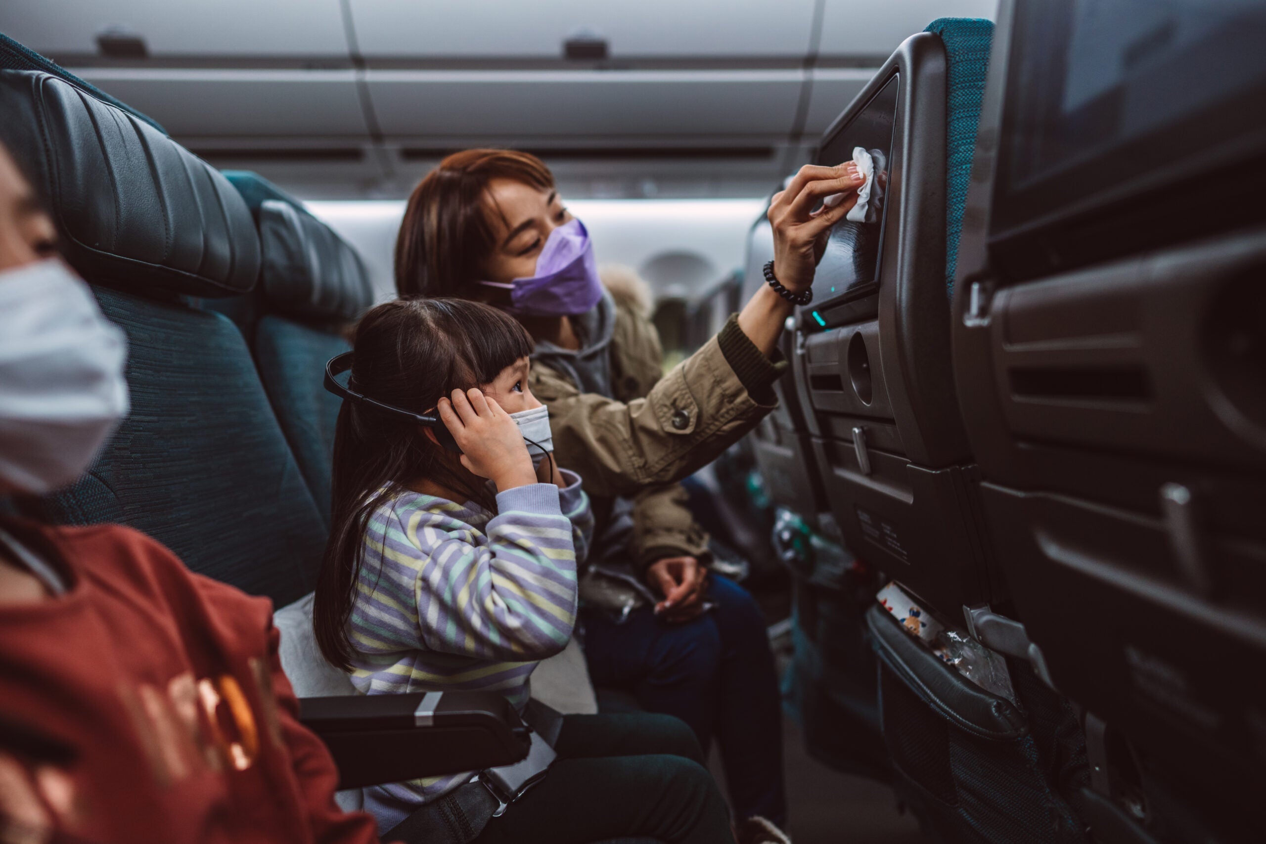 Mom & daughter in protective face masks cleaning the seat-back TV screen with disinfectant surface wipe while travelling on the airplane