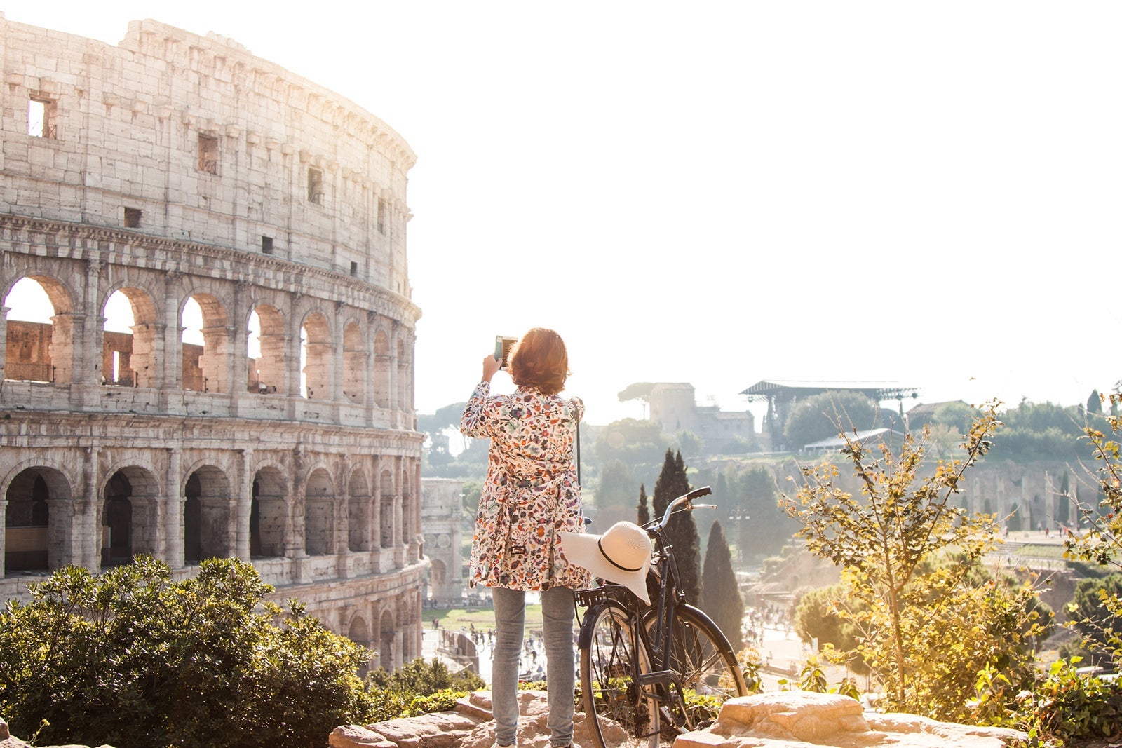 Beautiful young woman in colorful fashion dress alone on a hill with bike takes pictures of colosseum in Rome using smartphone camera at sunset. Attractive tourist girl with elegant straw hat
