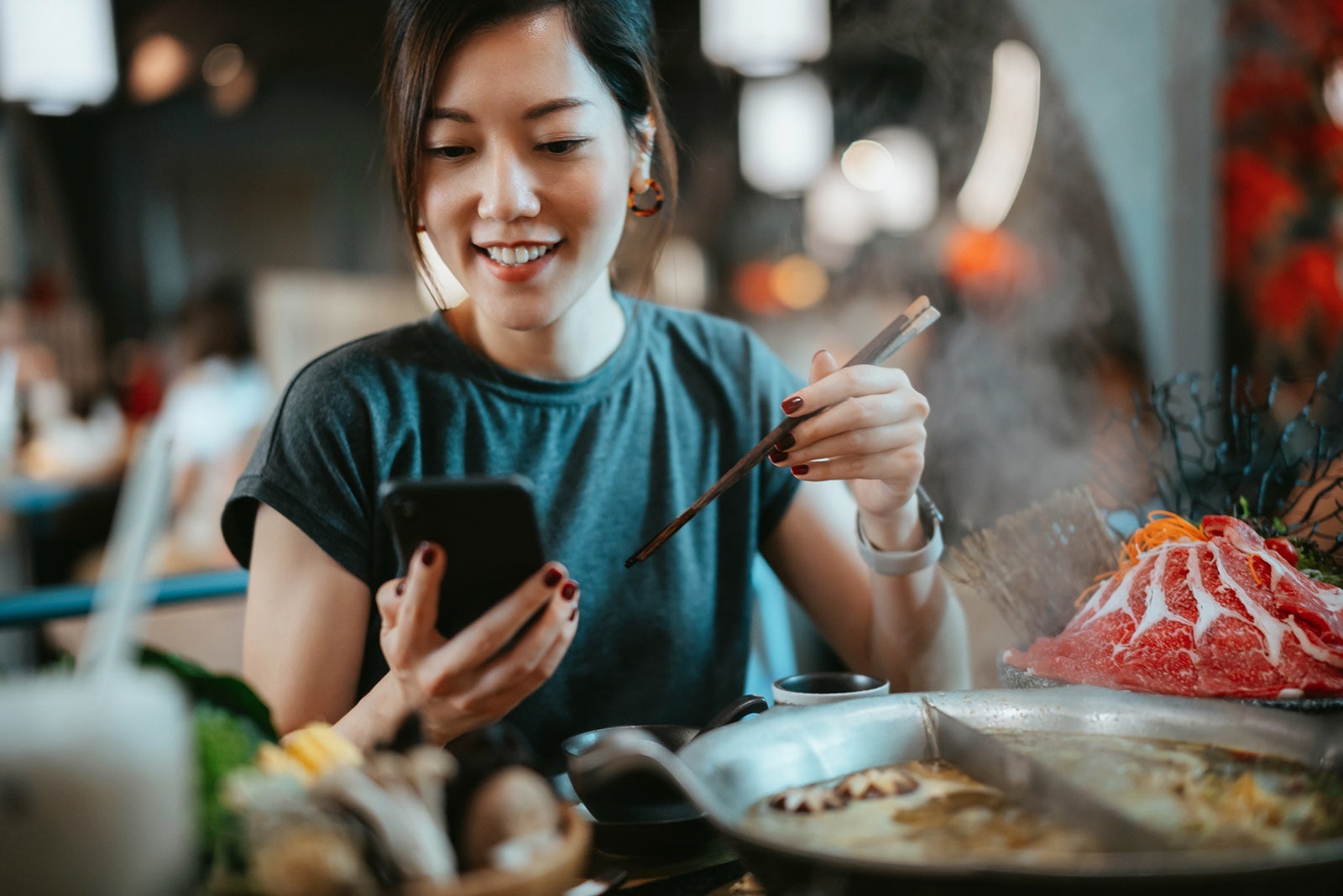 Beautiful smiling young Asian woman using smartphone while enjoying delicious traditional Chinese hotpot with assorted fresh ingredients in restaurant. Chinese cuisine and food. Eating out lifestyle. Technology in everyday life