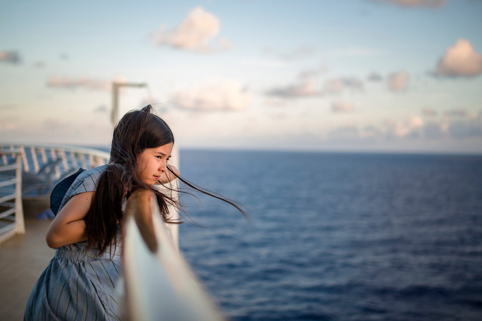 A young girl leans against a ship railing at sunset gazing at ocean