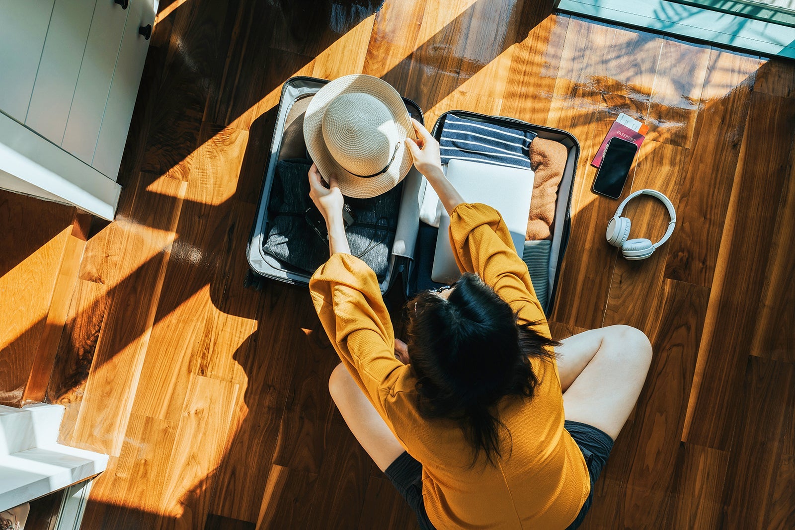 Overhead view of young Asian woman sitting on the floor in her bedroom, packing a suitcase for a trip. Getting ready for a vacation. Traveller's accessories. Travel and vacation concept