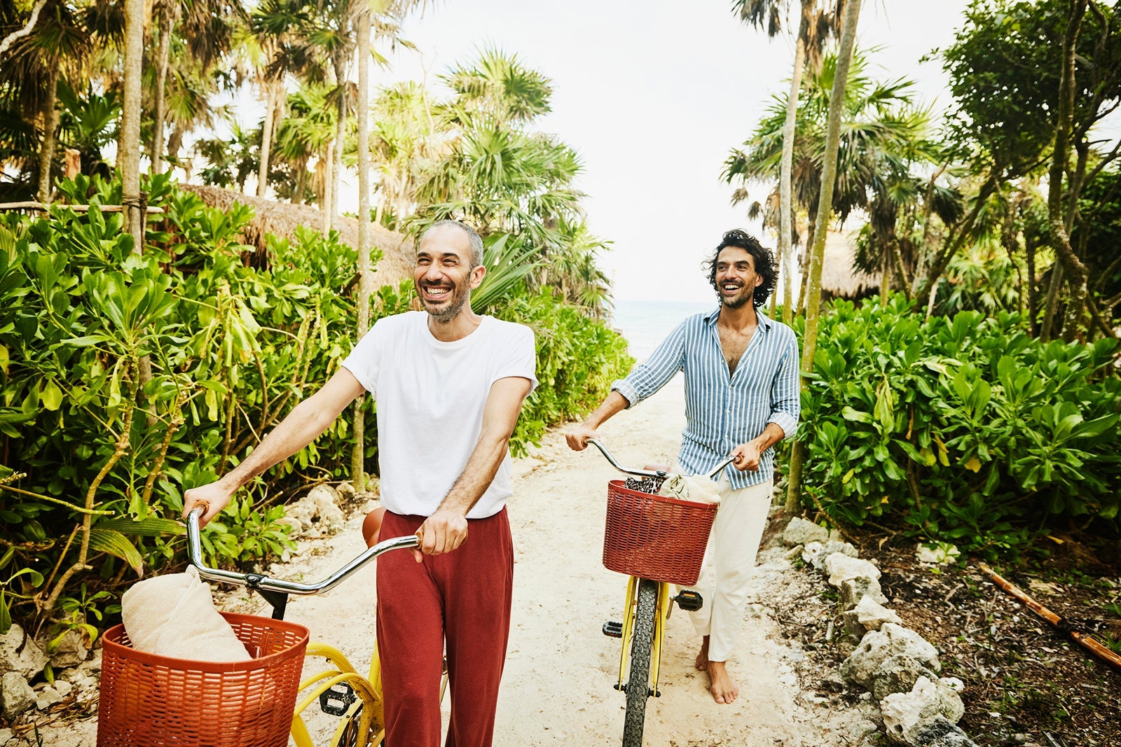 Medium wide shot of laughing gay couple walking bikes on path from beach at tropical resort