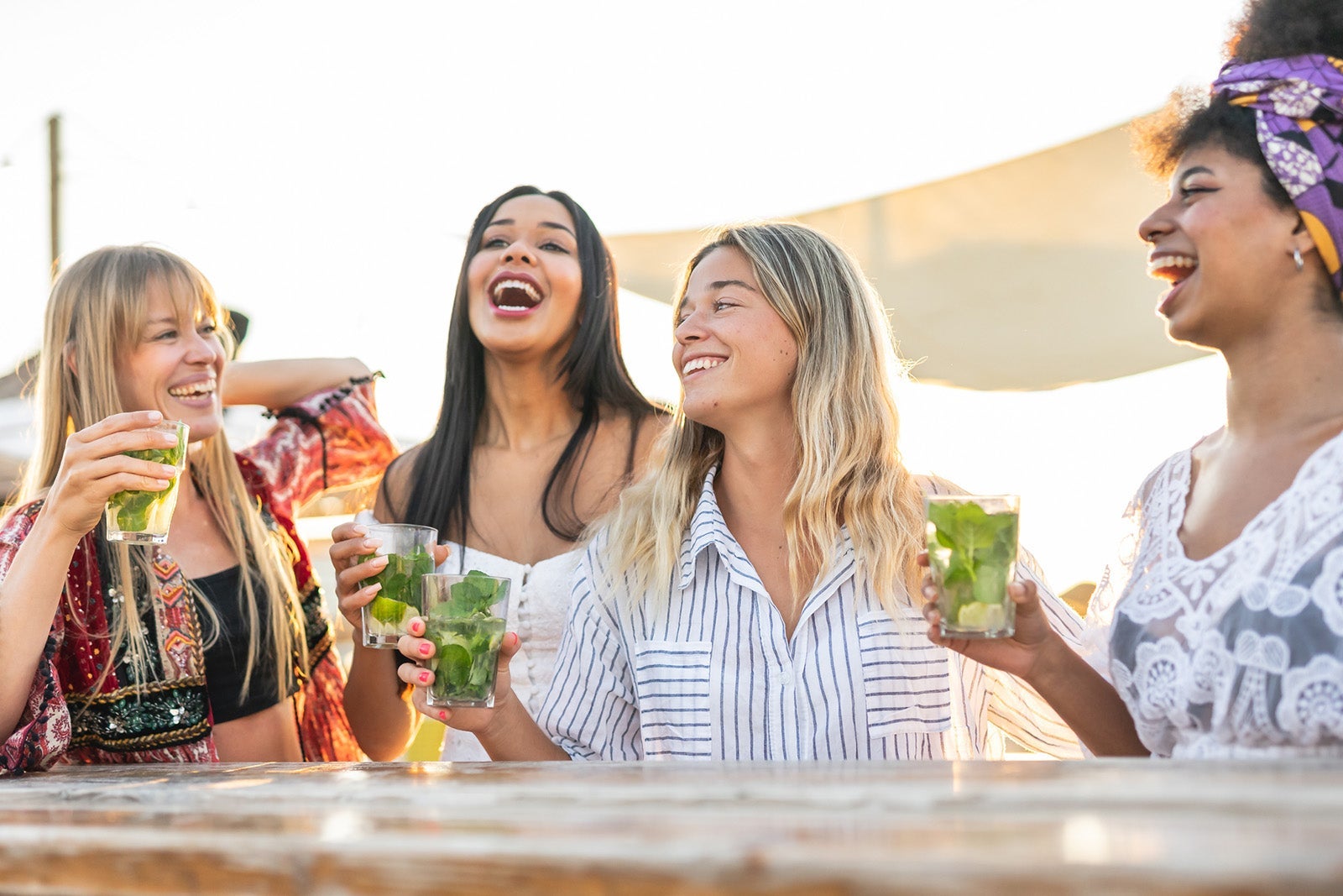 Young women toasting with mojito at chiringuito- Group of fashion girls celebrating bachelorette party while enjoying sunset and drinks on the beach