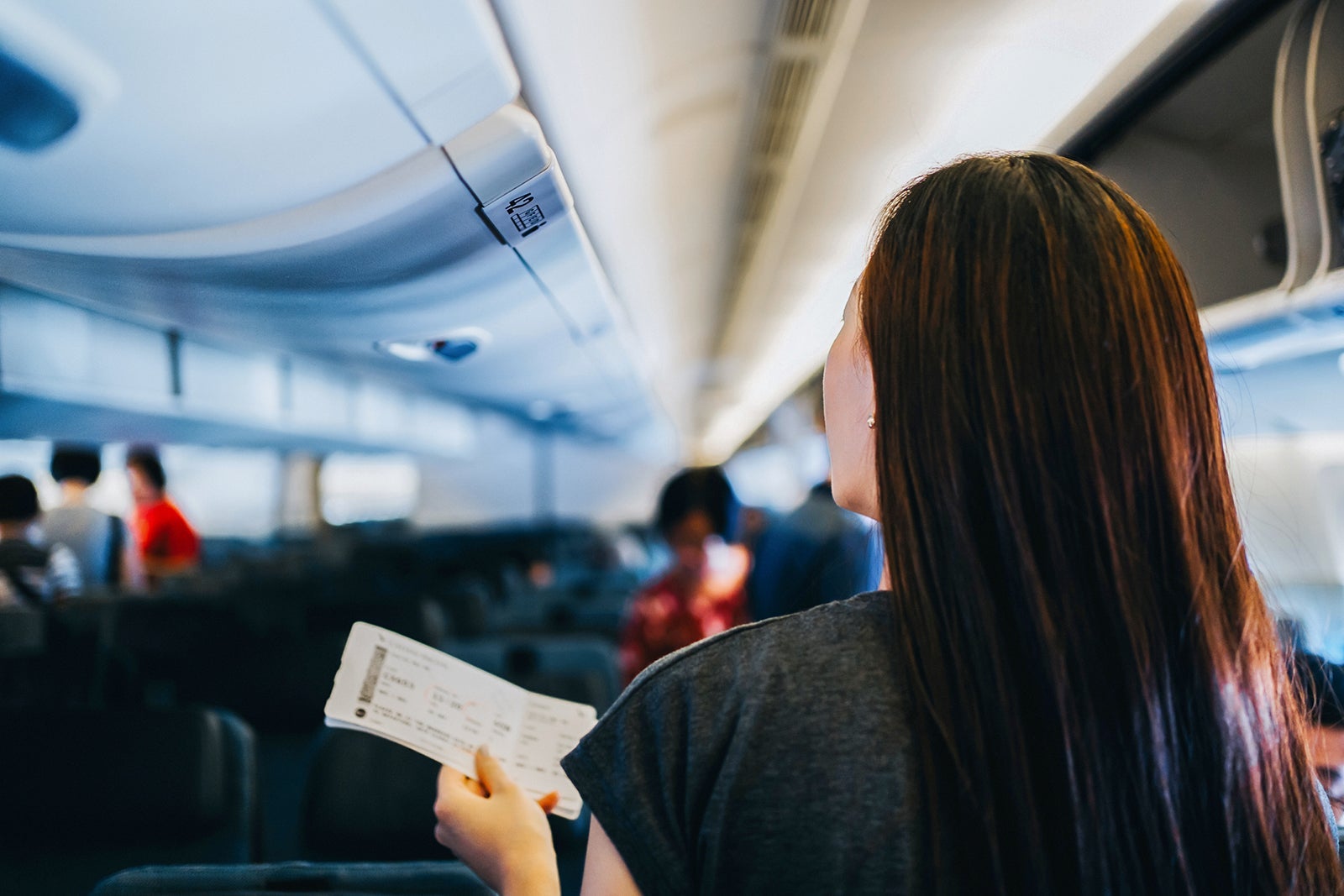 Rear view of young Asian female traveller holding her boarding pass, walking down the aisle in the airplane looking for her seat. Travel and vacation concept