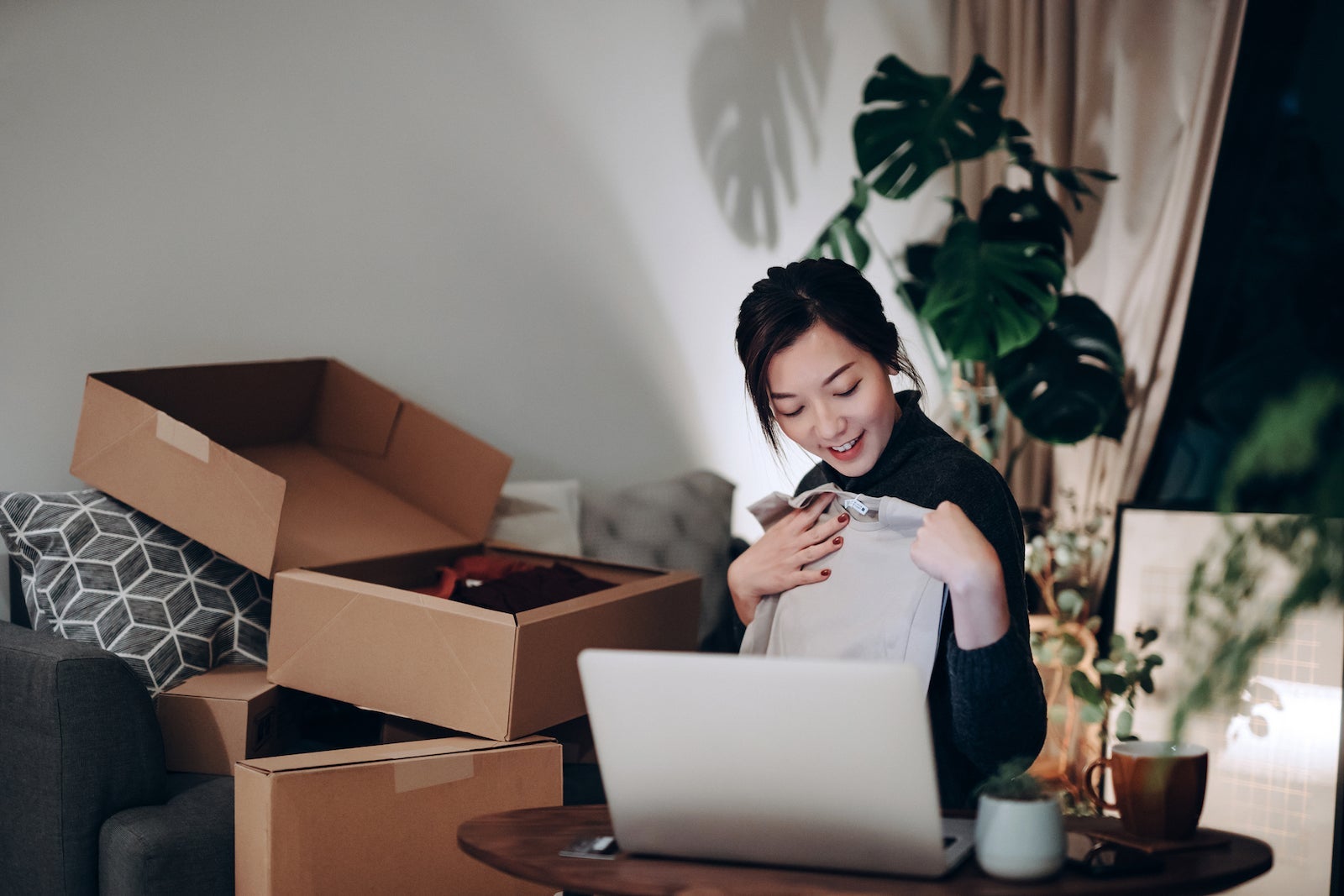 Young Asian woman unboxing new purchase clothings from cardboard box that received from her online shopping retail delivery at home. She is happy and excited to see the content from the box. Online shopping, trustworthy parcel delivery service