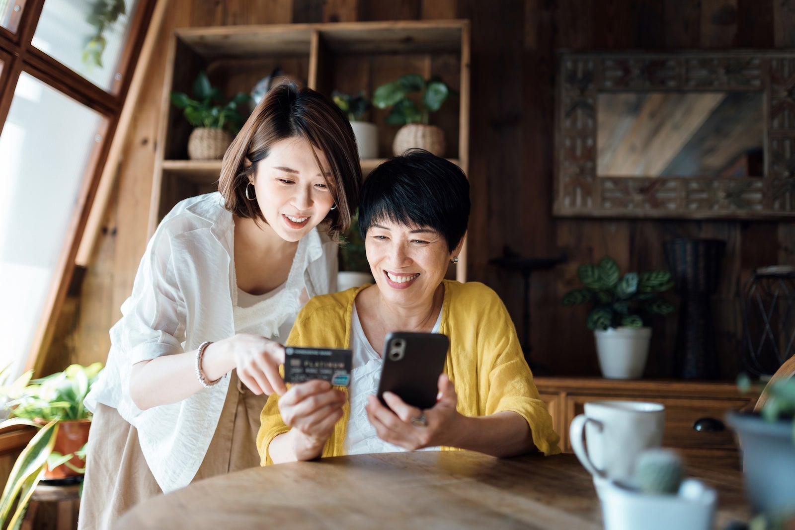Joyful Asian senior mother and daughter shopping online with smartphone together and making payment with credit card at home