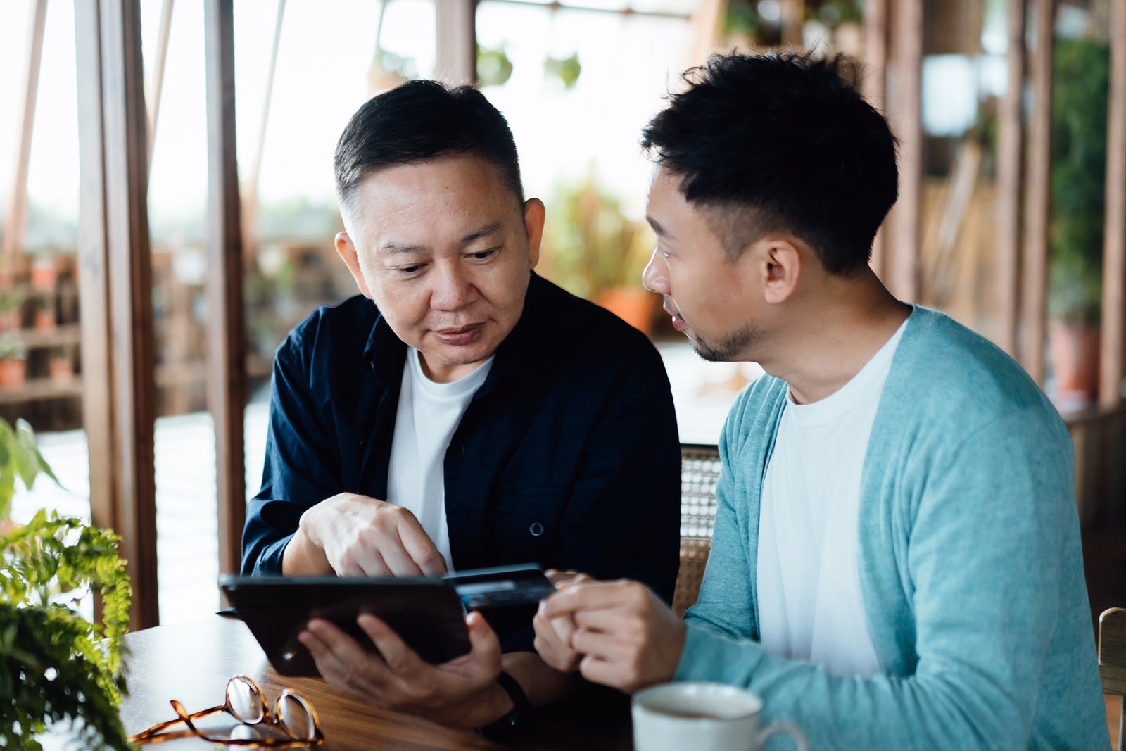 Asian son teaching his senior father how to shop online with a digital tablet and making payment with cred card at home. Engaging senior adults with technology