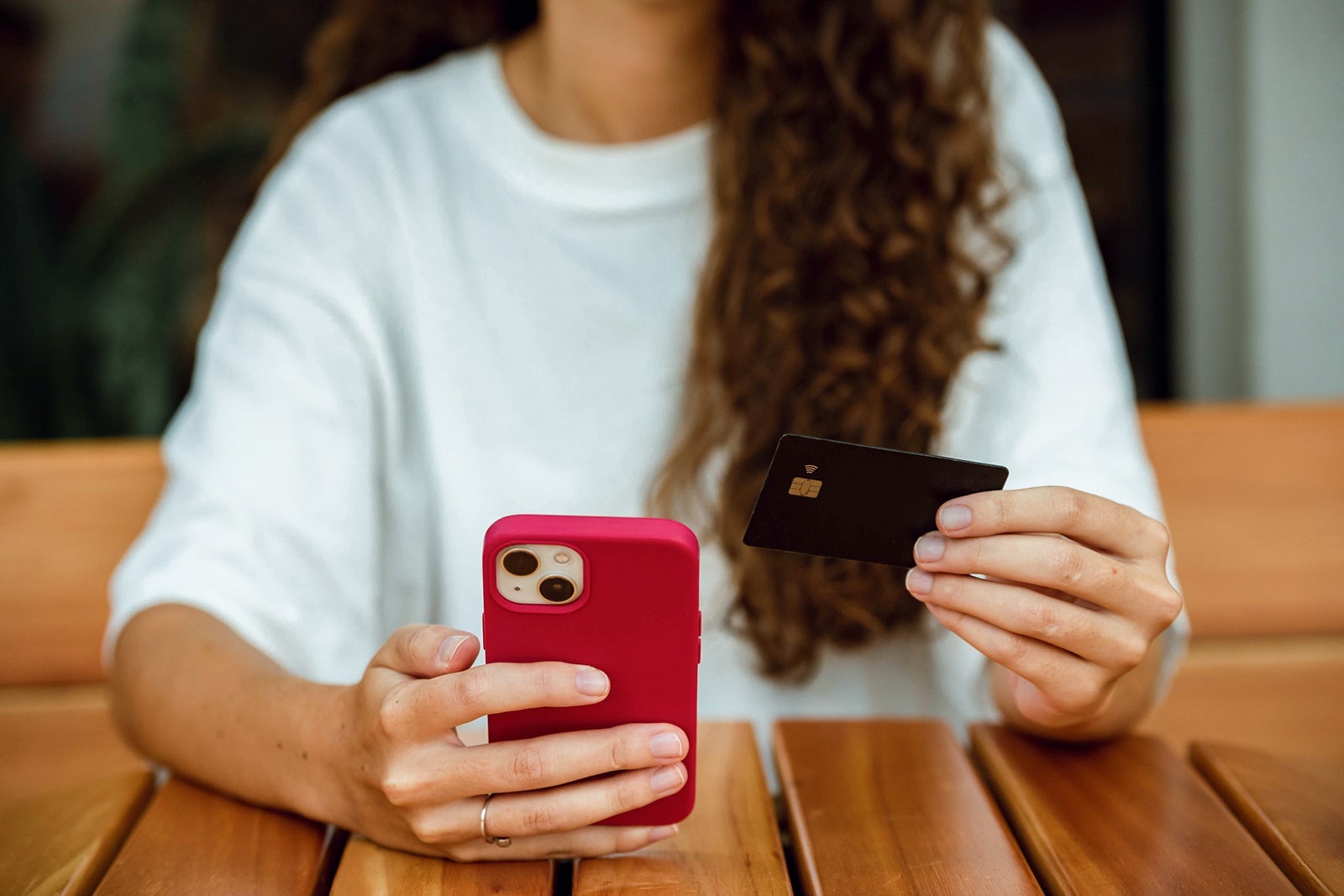Close op of a young adult woman's hand with long curly hair, wearing a white t-shirt, holding her cell phone with red cover and checking a black credit card