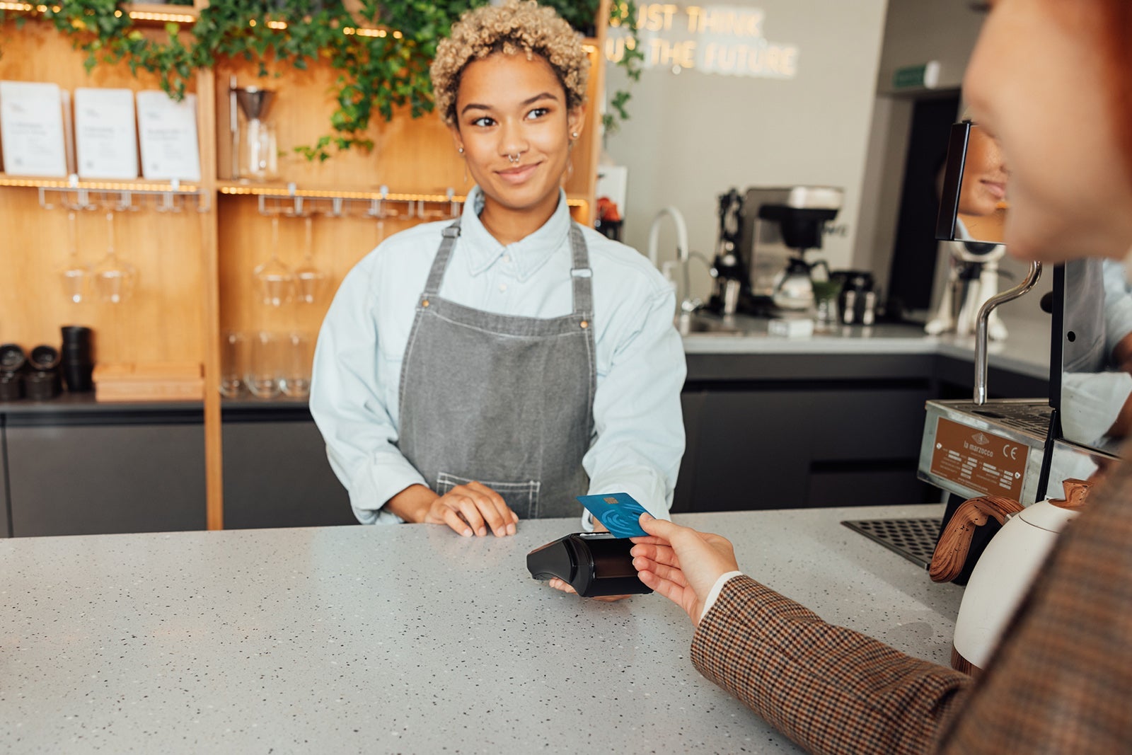 Woman bartender receives payment from a customer. Barista in apron holding pos terminal while customer paying buy card.