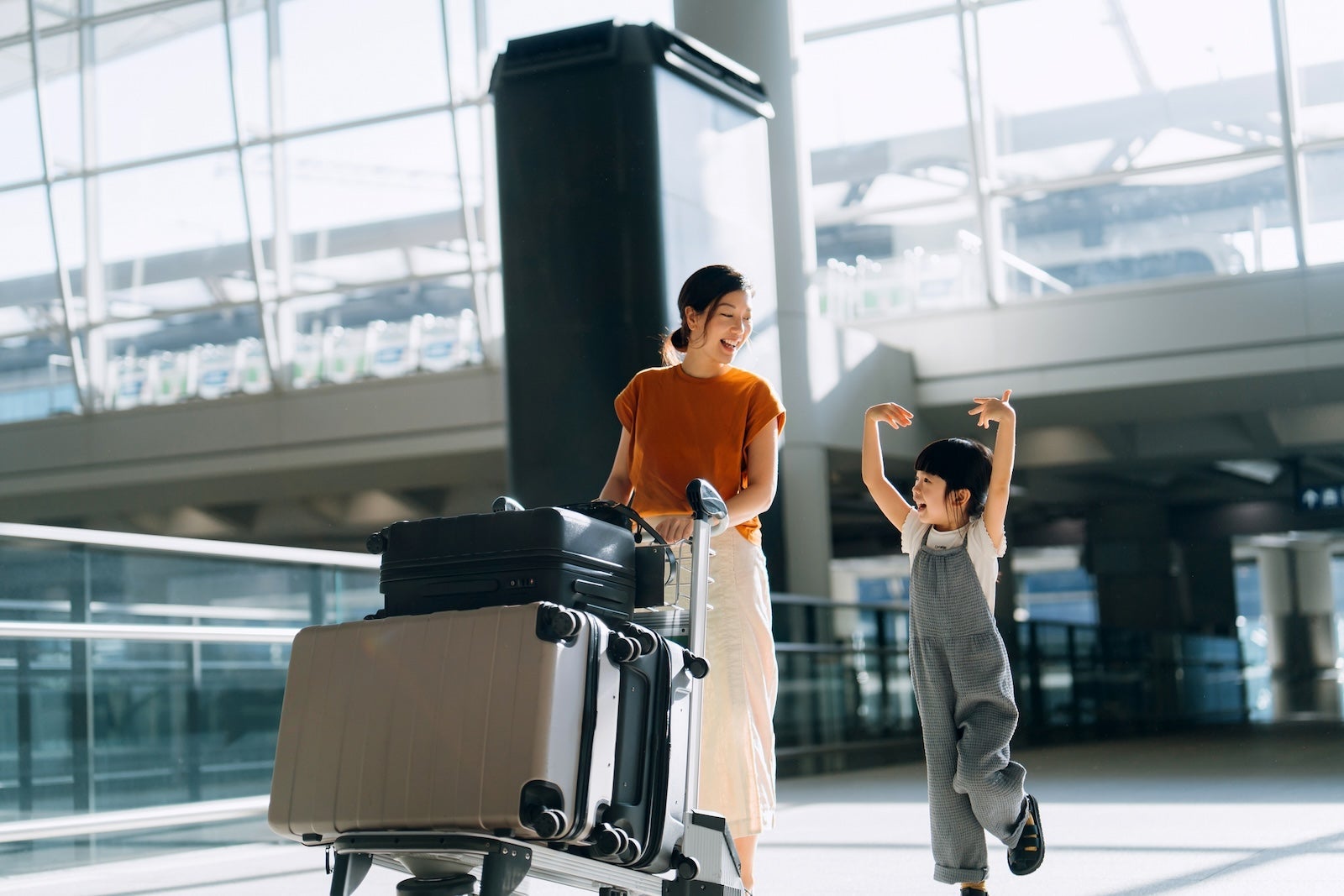 Joyful young Asian mother and little daughter travelling together, they are pushing a luggage trolley with suitcases at airport terminal. Ready for a trip. Travel and vacation concept