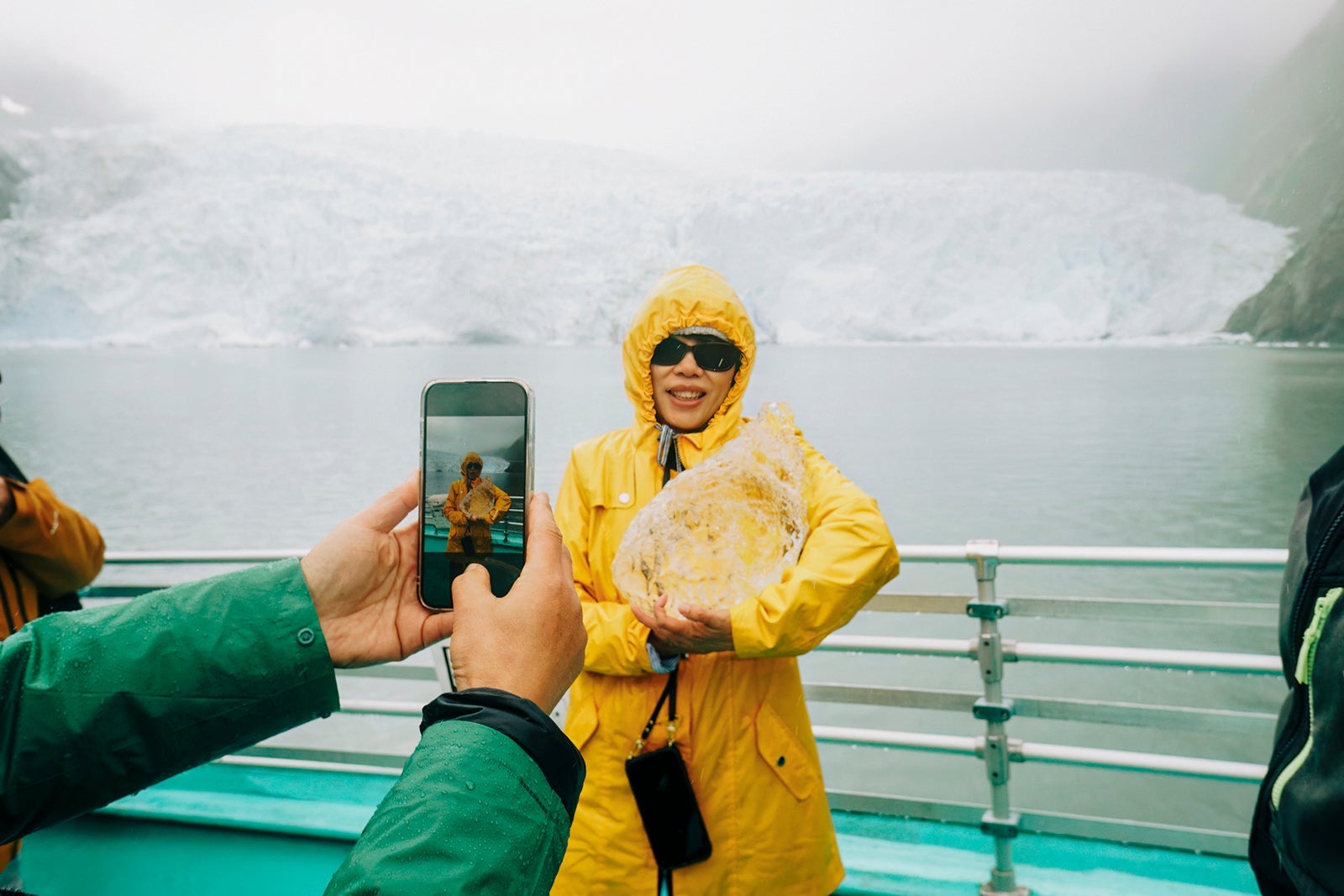Tourist taking smartphone photos of a woman holding ice from an iceberg. Holgate Glacier in the rain. Kenai Fjords National Park. Seward. Alaska.