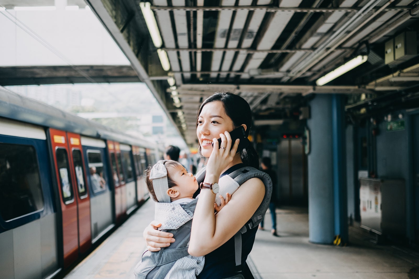 Busy young working mother with baby girl talking on smartphone while waiting for subway in subway station platform