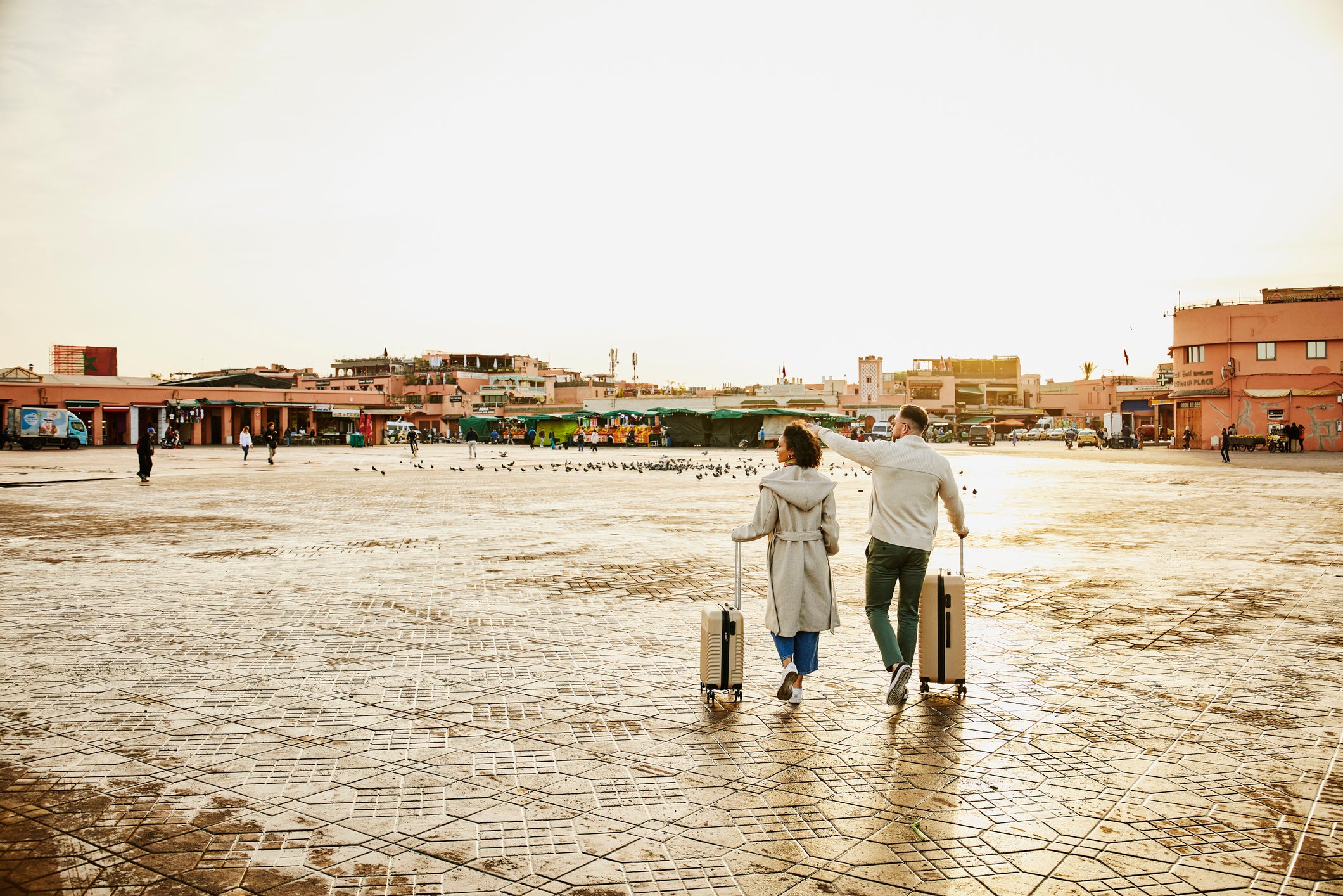 Wide shot rear view of couple with rolling luggage walking across Jemaa el-Fna square at sunrise during vacation in Marrakech